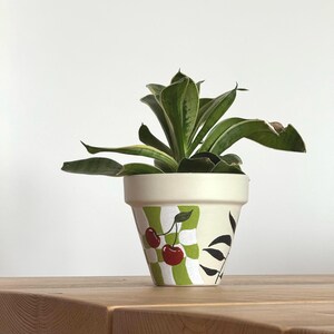 May include: A potted plant with green leaves sits in a cream-colored pot. The pot is decorated with a green and white checkered pattern, red cherries, and black leaf accents. The pot is on a wooden surface.