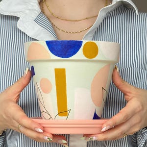 May include: A hand-painted ceramic plant pot with a matching saucer. The pot is off-white with abstract shapes in blue, yellow, and pink. A vertical gold rectangle and black line art are also featured. The saucer is light pink.