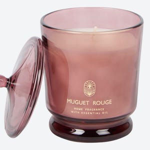 May include: A mauve-colored glass candle jar with a matching lid, featuring a white wax candle inside. The front of the glass displays a gold emblem above the text MUGUET ROUGE and the words HOME FRAGRANCE WITH ESSENTIAL OIL in gold lettering.