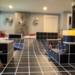 Peut inclure: Une scène de jeu de société avec des figurines miniatures sur une grille noire et blanche. Les figurines comprennent un soldat blindé blanc et gris et plusieurs figurines avec des sabres laser. Le décor est une pièce aux murs gris et aux éléments décoratifs.
