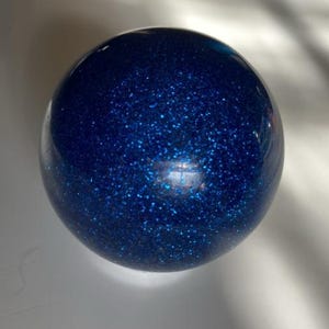 May include: A spherical object with a smooth, transparent surface containing a dense concentration of deep blue glitter particles that catch the light, creating a sparkling effect throughout the interior of the round, glass-like orb.