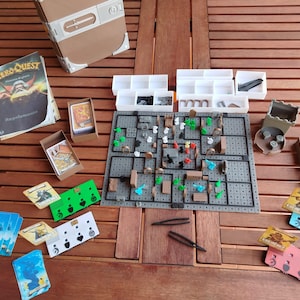 May include: A tabletop game setup featuring a game board, cards, miniature figures, dice, and storage boxes. The game board is gray with a grid pattern, and the cards have various illustrations. The game is set on a wooden table.