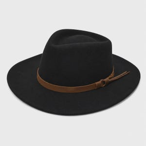Può includere: Un cappello fedora nero con tesa larga e corona pizzicata. Il cappello presenta una fascia in pelle marrone liscia avvolta attorno alla base, che include un piccolo dettaglio a nodo e due sottili strisce di pelle che sporgono dal lato.