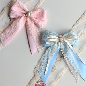 May include: Two decorative bows. One is light pink with lace trim and a pearl-centered embellishment. The other is light blue with cream-colored ribbon and a cameo-style center. Both have long, flowing tails.