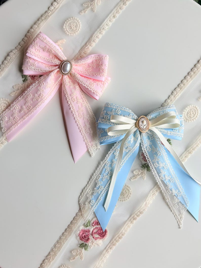 May include: Two decorative bows. One is light pink with lace trim and a pearl-centered embellishment. The other is light blue with cream-colored ribbon and a cameo-style center. Both have long, flowing tails.