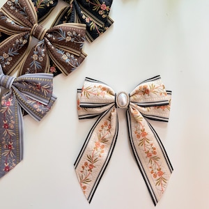 May include: Assortment of decorative hair bows in various colors and patterns. The bows feature floral designs, stripes, and a pearl-like embellishment. Colors include black, brown, gold, and gray. The bows are arranged on a white surface.