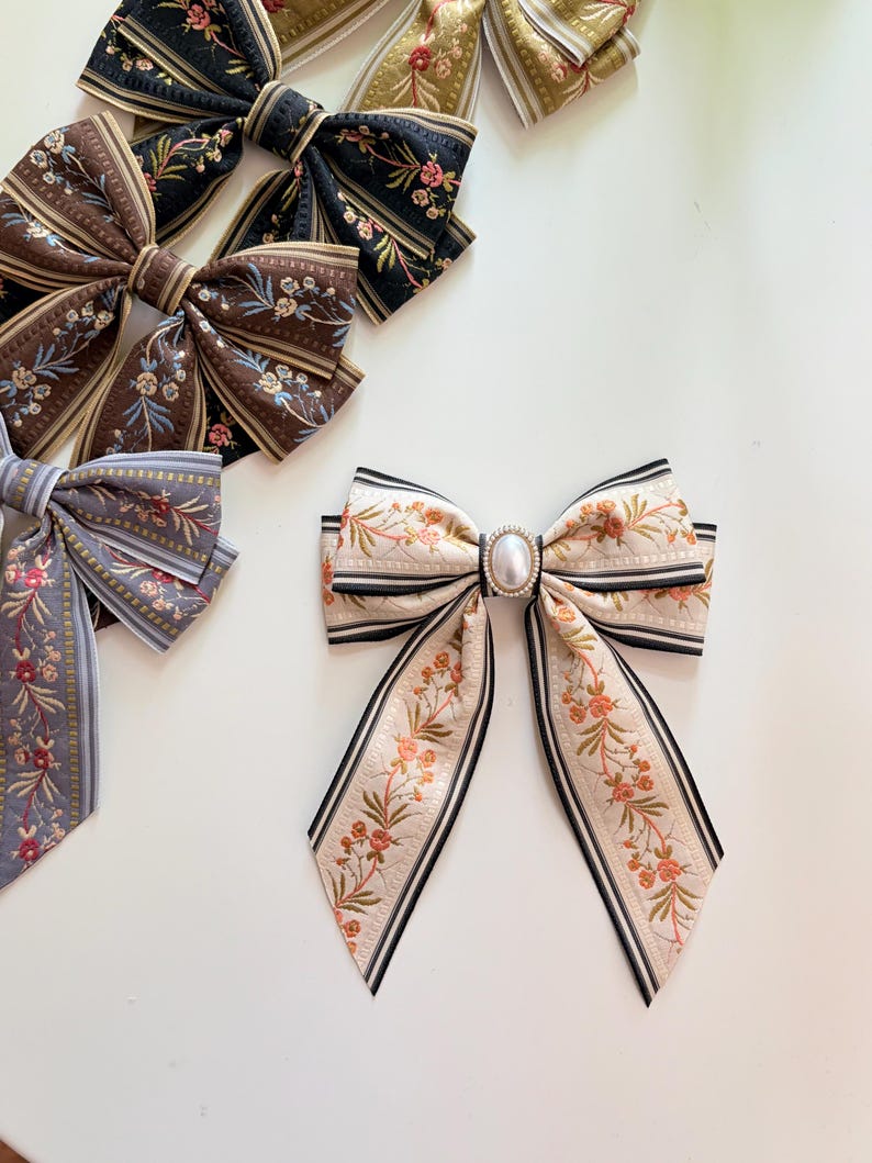 May include: Assortment of decorative hair bows in various colors and patterns. The bows feature floral designs, stripes, and a pearl-like embellishment. Colors include black, brown, gold, and gray. The bows are arranged on a white surface.