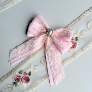 May include: A light pink bow with a pearl-like center, surrounded by a silver-colored setting. The bow is made of a lace-trimmed ribbon. Floral embroidered lace trim is visible in the background.