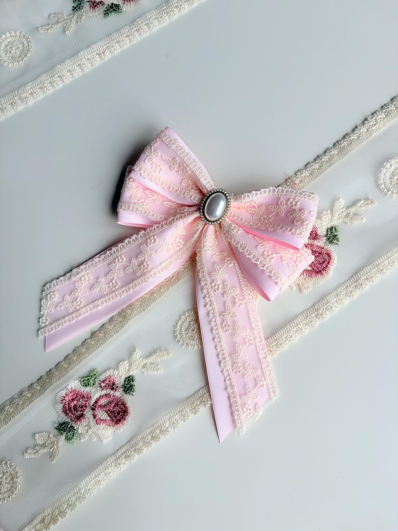 May include: A light pink bow with a pearl-like center, surrounded by a silver-colored setting. The bow is made of a lace-trimmed ribbon. Floral embroidered lace trim is visible in the background.