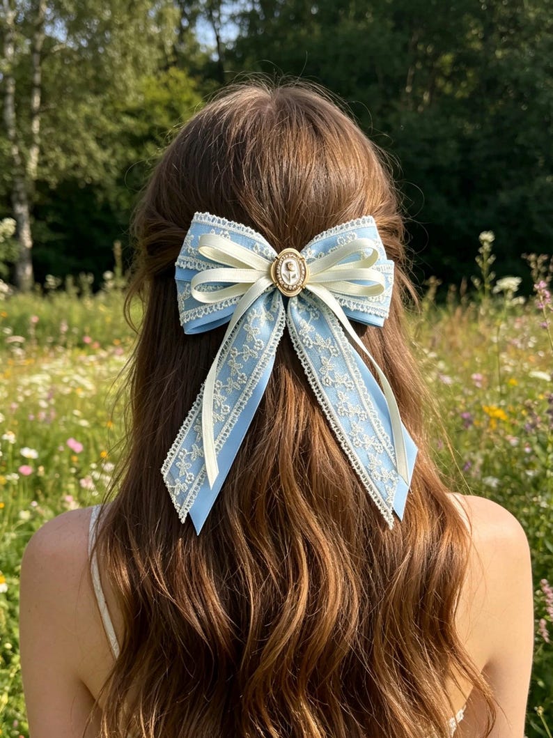 May include: A light blue hair bow with ivory accents and delicate lace trim. The bow features a small, ornate gold-toned centerpiece. The hair accessory is set against a backdrop of long, brown hair and a field of wildflowers.