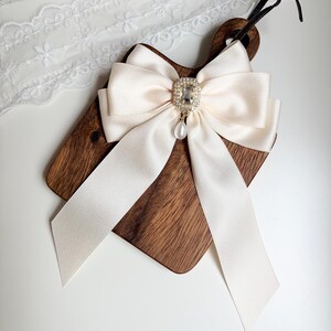 May include: A cream-colored satin bow with long, flowing tails is centered on a small, dark wooden cutting board. The bow features a sparkling, rectangular rhinestone centerpiece with a dangling pearl. A black cord is attached to the board.