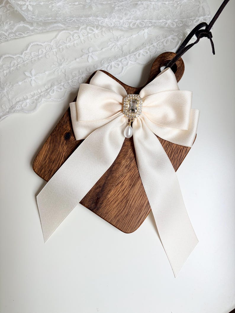 May include: A cream-colored satin bow with long, flowing tails is centered on a small, dark wooden cutting board. The bow features a sparkling, rectangular rhinestone centerpiece with a dangling pearl. A black cord is attached to the board.