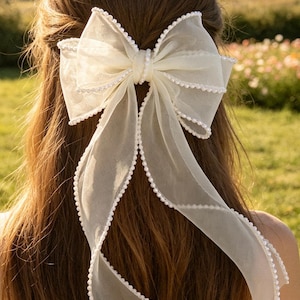 Op de afbeelding: Een grote, gebroken witte haarsstrik van transparante organza met een delicate rand van parelachtige kraaltjes langs alle zijden. De strik is opgebouwd uit meerdere lagen doorschijnende stof, wat zorgt voor een volumineuze vorm met twee lange slierten.