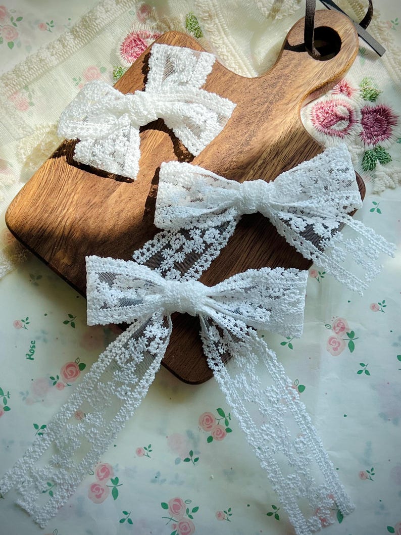 Puede incluir: Tres lazos de encaje blanco de diferentes tama&ntilde;os est&aacute;n dispuestos sobre una tabla de cortar de madera. Los lazos tienen intrincados patrones florales y largas colas fluidas. El fondo incluye una tela con estampado floral.