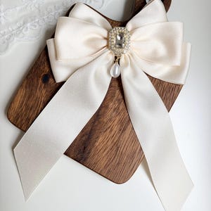 May include: Ivory satin bow with a sparkling rhinestone centerpiece and a dangling pearl pendant. The bow is attached to a wooden cutting board. The bow's long, flowing ribbons add a touch of elegance.