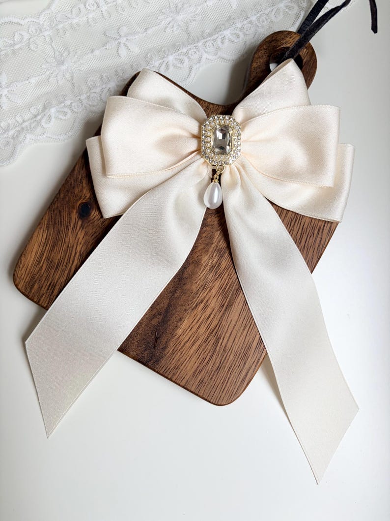May include: Ivory satin bow with a sparkling rhinestone centerpiece and a dangling pearl pendant. The bow is attached to a wooden cutting board. The bow's long, flowing ribbons add a touch of elegance.