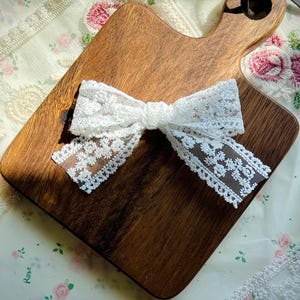 Puede incluir: Un lazo de encaje blanco est&aacute; centrado en una tabla de cortar de madera. El lazo est&aacute; hecho de encaje delicado con estampado floral. La tabla de cortar es marr&oacute;n y tiene un asa. El fondo es una tela estampada floral.