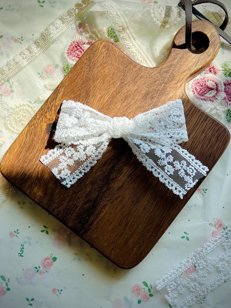 Puede incluir: Un lazo de encaje blanco est&aacute; centrado en una tabla de cortar de madera. El lazo est&aacute; hecho de encaje delicado con estampado floral. La tabla de cortar es marr&oacute;n y tiene un asa. El fondo es una tela estampada floral.