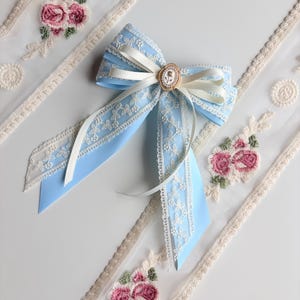 May include: A light blue bow with white lace trim and a small gold-colored embellishment. The bow is paired with white ribbon and sits near embroidered floral lace trim with pink roses.