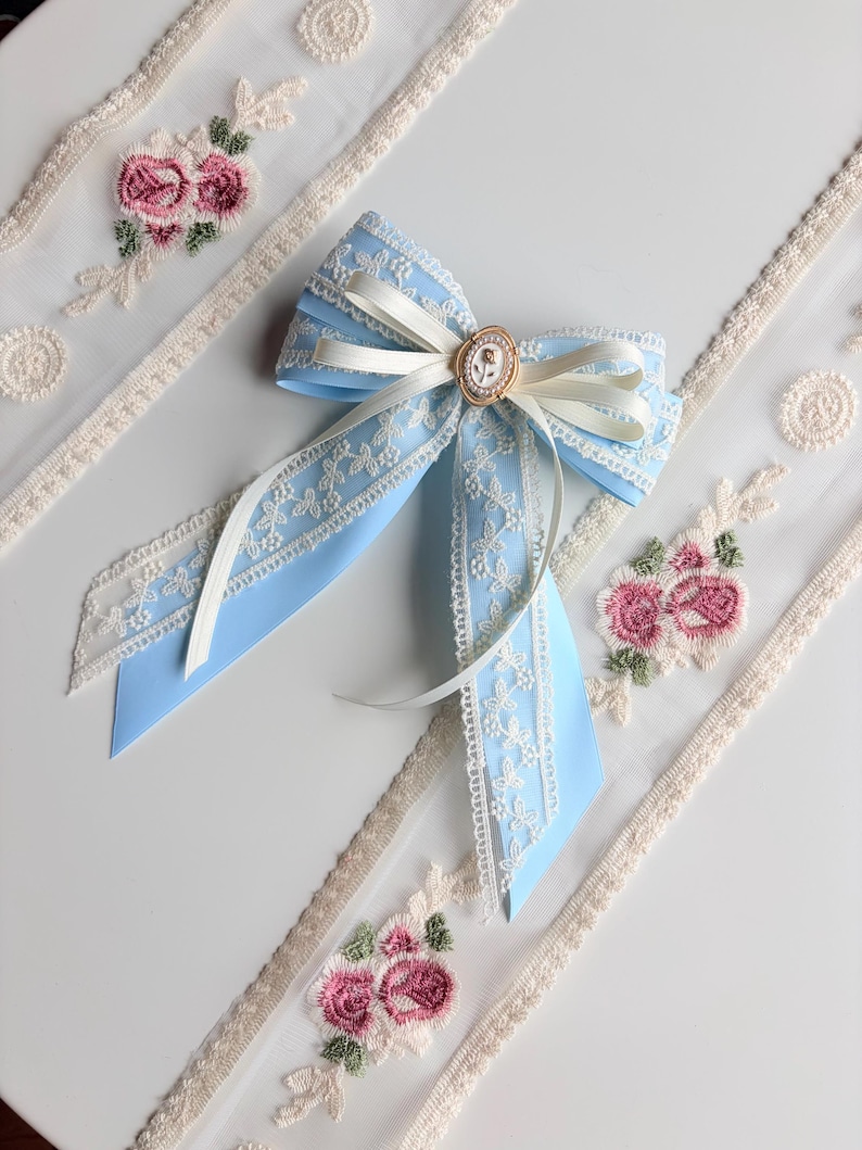 May include: A light blue bow with white lace trim and a small gold-colored embellishment. The bow is paired with white ribbon and sits near embroidered floral lace trim with pink roses.