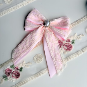 May include: A light pink bow with cream-colored lace trim and a pearl-like center. The bow is set against a white surface with embroidered floral and lace details, creating a delicate and feminine aesthetic.
