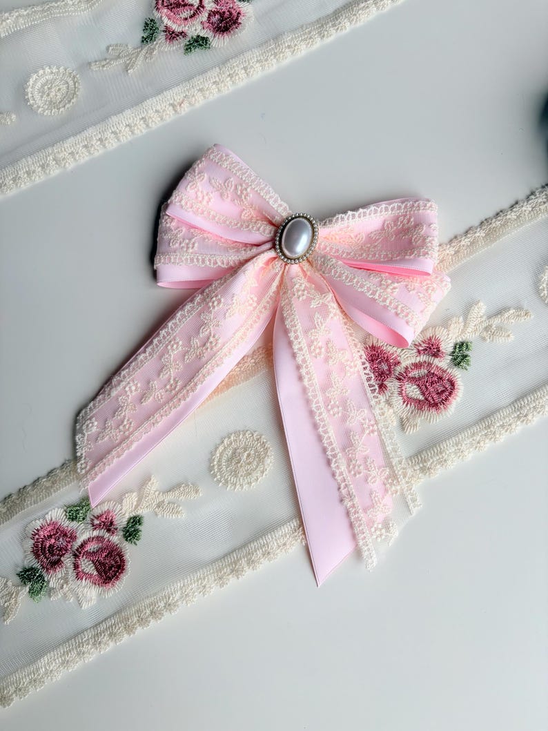 May include: A light pink bow with cream-colored lace trim and a pearl-like center. The bow is set against a white surface with embroidered floral and lace details, creating a delicate and feminine aesthetic.