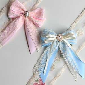 May include: Two decorative bows, one pink and one light blue, with lace trim and a central embellishment. The pink bow has a pearl-like center, while the blue bow features a cameo-style center. Both bows have long, flowing ribbons.