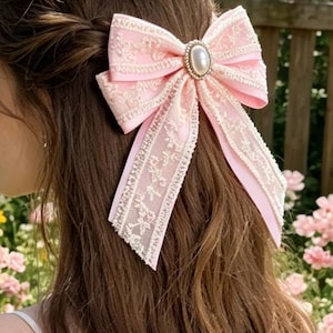 May include: A pink hair bow with a delicate lace overlay and a pearl-like center embellishment. The bow is tied and attached to a person's hair. The background features a wooden fence and pink flowers, suggesting an outdoor setting.