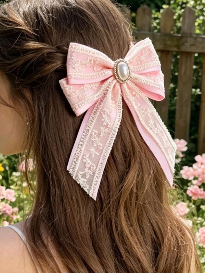 May include: A pink hair bow with a delicate lace overlay and a pearl-like center embellishment. The bow is tied and attached to a person's hair. The background features a wooden fence and pink flowers, suggesting an outdoor setting.