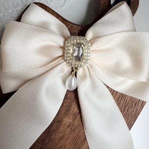 May include: Cream-colored satin bow with a rectangular rhinestone and pearl pendant. The bow is centered on a dark wooden surface. The bow's ribbon is wide and has a slight sheen.