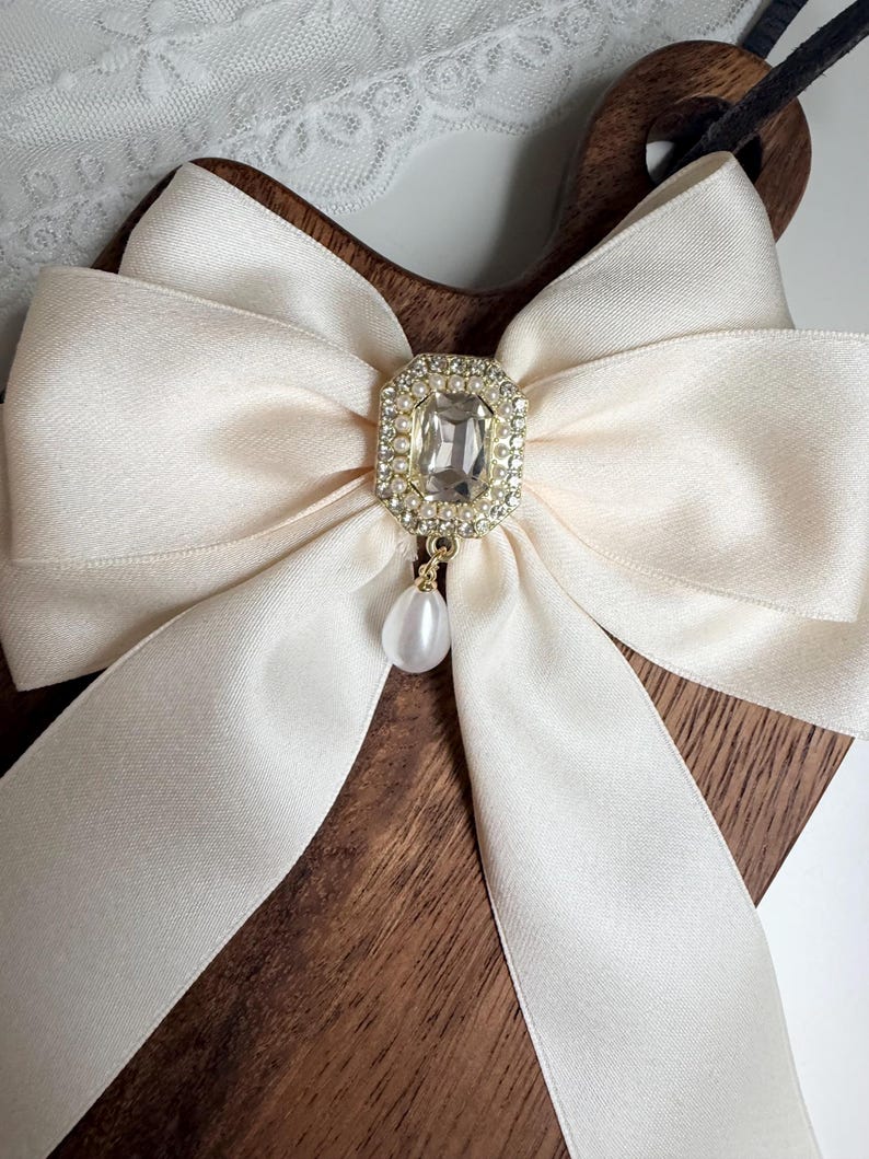 May include: Cream-colored satin bow with a rectangular rhinestone and pearl pendant. The bow is centered on a dark wooden surface. The bow's ribbon is wide and has a slight sheen.