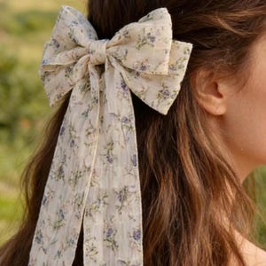 Könnte beinhalten: Eine große, cremefarbene Haarschleife mit einem floralen Muster aus blauen und grünen Blumen. Die Schleife ist im Haar gebunden, mit langen, fließenden Bändern. Das Accessoire verleiht jeder Frisur einen Hauch von Eleganz und Charme.