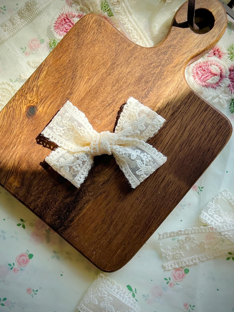 Puede incluir: Un lazo de encaje color crema est&aacute; centrado en una tabla de cortar de madera. El lazo est&aacute; hecho de encaje delicado y estampado. La tabla de cortar es de color marr&oacute;n c&aacute;lido y tiene un asa. El fondo es una tela con estampado floral.