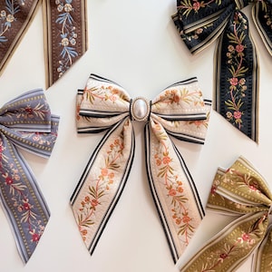 May include: An assortment of decorative hair bows with floral patterns. The bows feature various colors, including cream, black, brown, and olive green, with delicate floral embroidery and pearl accents. The bows are arranged on a white surface.