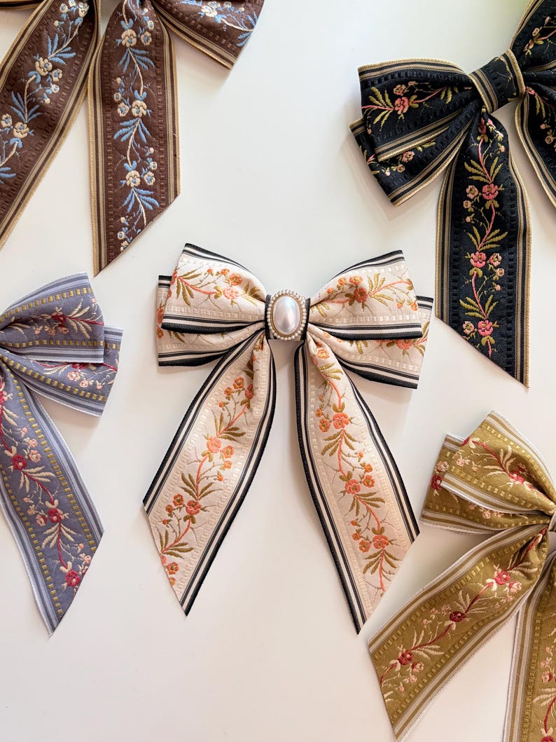 May include: An assortment of decorative hair bows with floral patterns. The bows feature various colors, including cream, black, brown, and olive green, with delicate floral embroidery and pearl accents. The bows are arranged on a white surface.