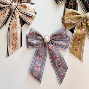 May include: Several decorative hair bows in various colors and patterns are displayed on a white surface. The bows feature floral embroidery, striped designs, and a pearl-like embellishment. Colors include cream, brown, black, gold, and lavender.
