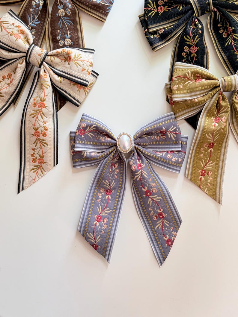 May include: Several decorative hair bows in various colors and patterns are displayed on a white surface. The bows feature floral embroidery, striped designs, and a pearl-like embellishment. Colors include cream, brown, black, gold, and lavender.