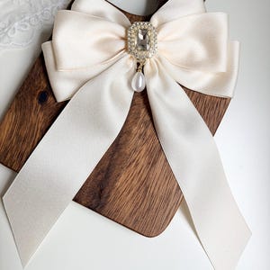May include: Ivory satin bow with a rhinestone and pearl embellishment. The bow is centered on a wooden cutting board. The bow's long, flowing ribbons and the sparkling accents create an elegant, feminine accessory.