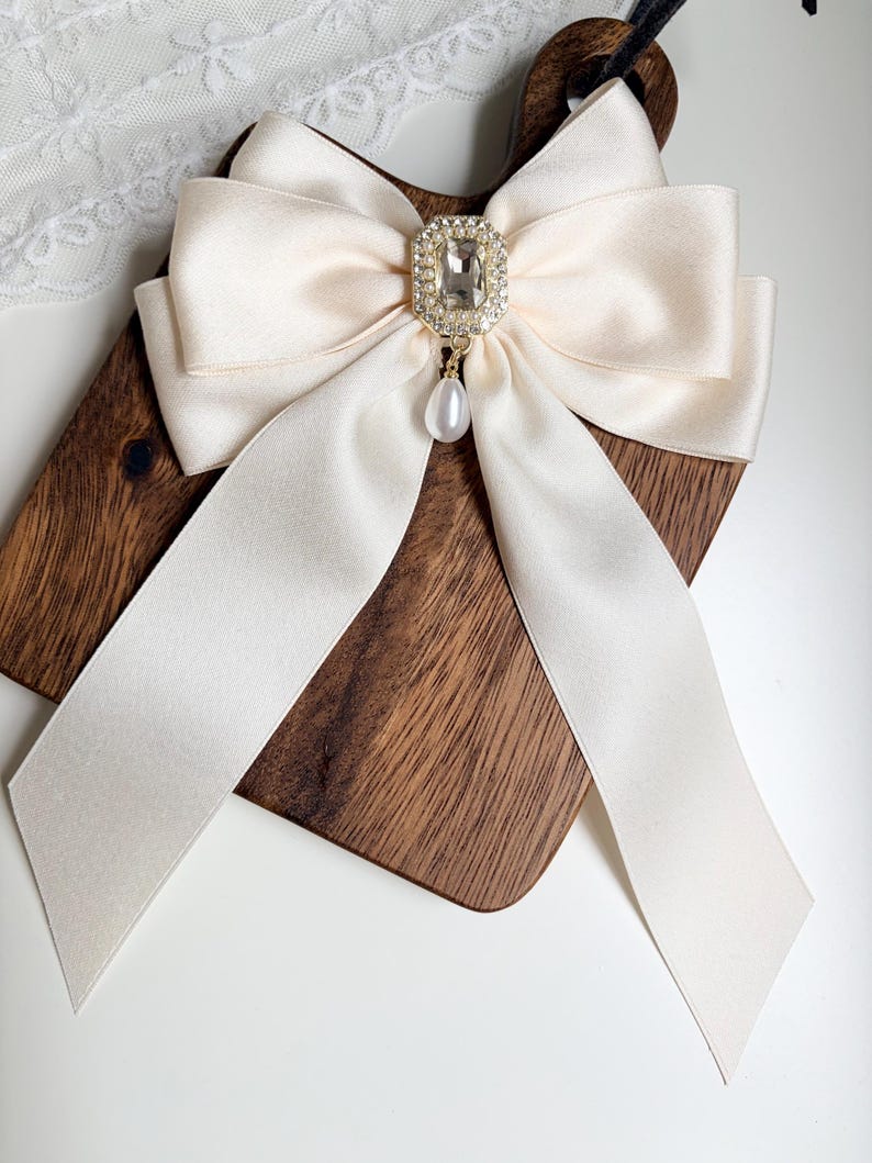 May include: Ivory satin bow with a rhinestone and pearl embellishment. The bow is centered on a wooden cutting board. The bow's long, flowing ribbons and the sparkling accents create an elegant, feminine accessory.