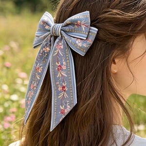 May include: A blue hair bow with a floral pattern is secured in the hair. The bow features a light blue background with vertical stripes and a floral design in pink and green. The bow is tied in a classic bow shape with long, flowing tails.