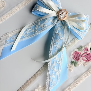 May include: A light blue bow with white lace trim and a gold-colored oval centerpiece. The bow is adorned with a white ribbon and is surrounded by embroidered floral lace trim.