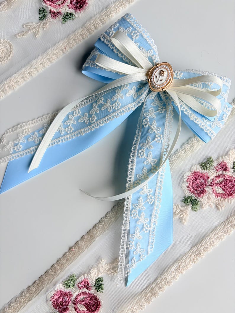 May include: A light blue bow with white lace trim and a gold-colored oval centerpiece. The bow is adorned with a white ribbon and is surrounded by embroidered floral lace trim.