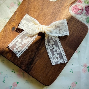 Puede incluir: Lazo de encaje color marfil sobre una tabla de cortar de madera. El lazo presenta un patr&oacute;n floral y est&aacute; atado en el centro. La tabla de cortar es marr&oacute;n con un asa. El fondo es una tela con estampado floral.