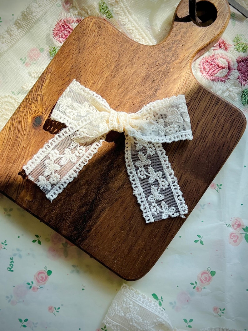 Puede incluir: Lazo de encaje color marfil sobre una tabla de cortar de madera. El lazo presenta un patr&oacute;n floral y est&aacute; atado en el centro. La tabla de cortar es marr&oacute;n con un asa. El fondo es una tela con estampado floral.