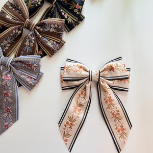 May include: Several fabric hair bows in various colors and patterns are displayed on a white surface. The bows feature floral designs and striped accents. Colors include black, brown, gray, and cream with pink and green floral details.