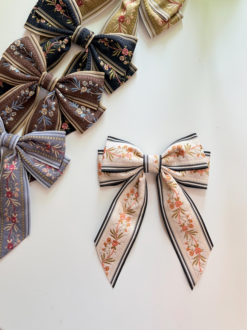 May include: Several fabric hair bows in various colors and patterns are displayed on a white surface. The bows feature floral designs and striped accents. Colors include black, brown, gray, and cream with pink and green floral details.