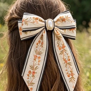 May include: A cream-colored hair bow with a floral pattern in orange and green, accented with black stripes. The bow features a pearl-like center and long, flowing tails. The accessory is set against a backdrop of brown hair and a blurred green background.