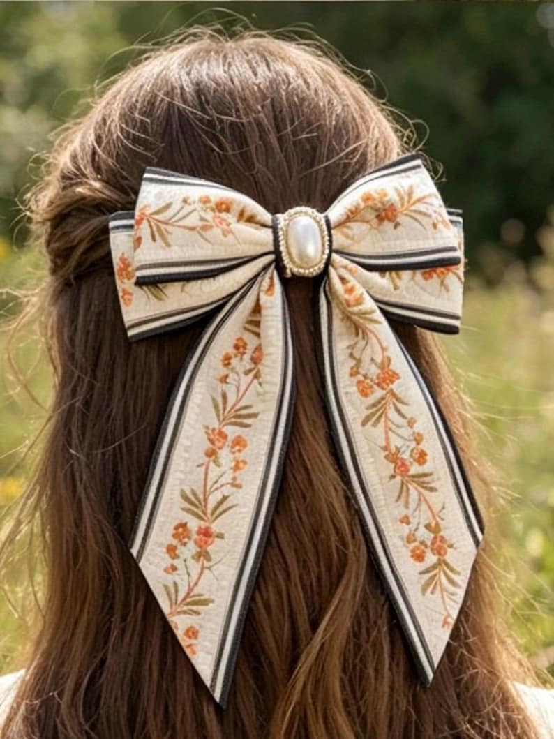 May include: A cream-colored hair bow with a floral pattern in orange and green, accented with black stripes. The bow features a pearl-like center and long, flowing tails. The accessory is set against a backdrop of brown hair and a blurred green background.