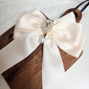 May include: A cream-colored satin bow with a rhinestone and pearl pendant. The bow is attached to a wooden paddle with a black handle. The bow's center features a square rhinestone piece surrounded by smaller rhinestones, with a dangling pearl.