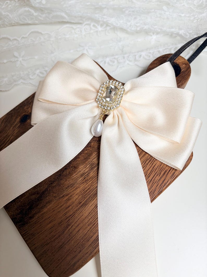 May include: A cream-colored satin bow with a rhinestone and pearl pendant. The bow is attached to a wooden paddle with a black handle. The bow's center features a square rhinestone piece surrounded by smaller rhinestones, with a dangling pearl.